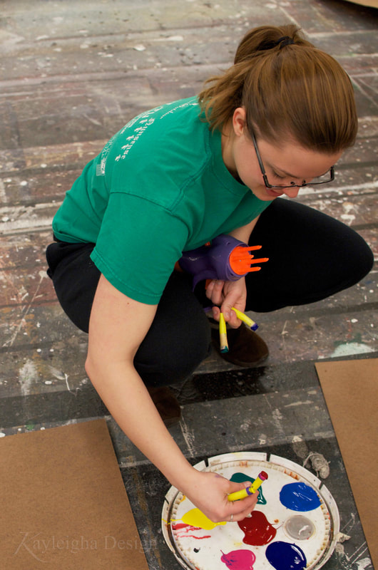 A light-haired woman with a ponytail wearing a green shirt and black sweats smiles as she dips yellow nerf gun darks into paint which is poured onto the lid of a white bucket to create a painter's palette.