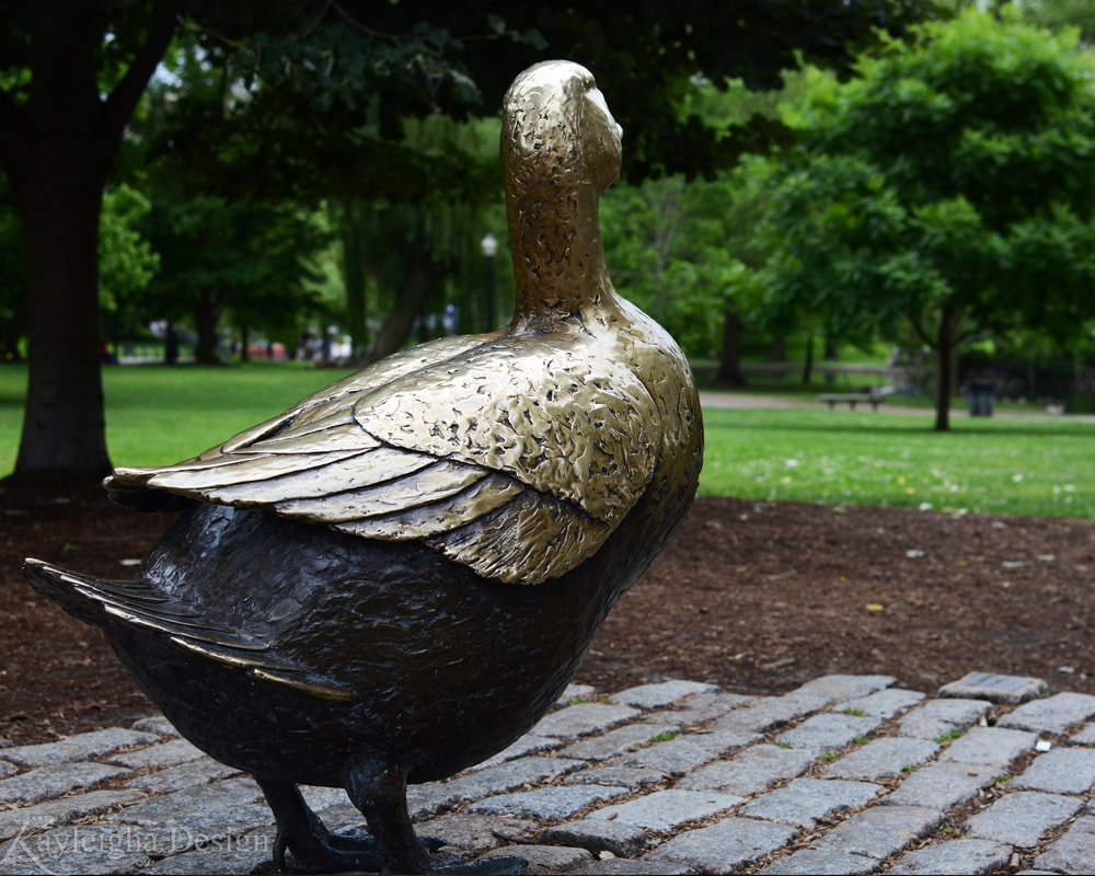 A color photo taken from behind a golden/brass colored sculpture of the mother duck. Taken as though the photographer is looking over her shoulder.