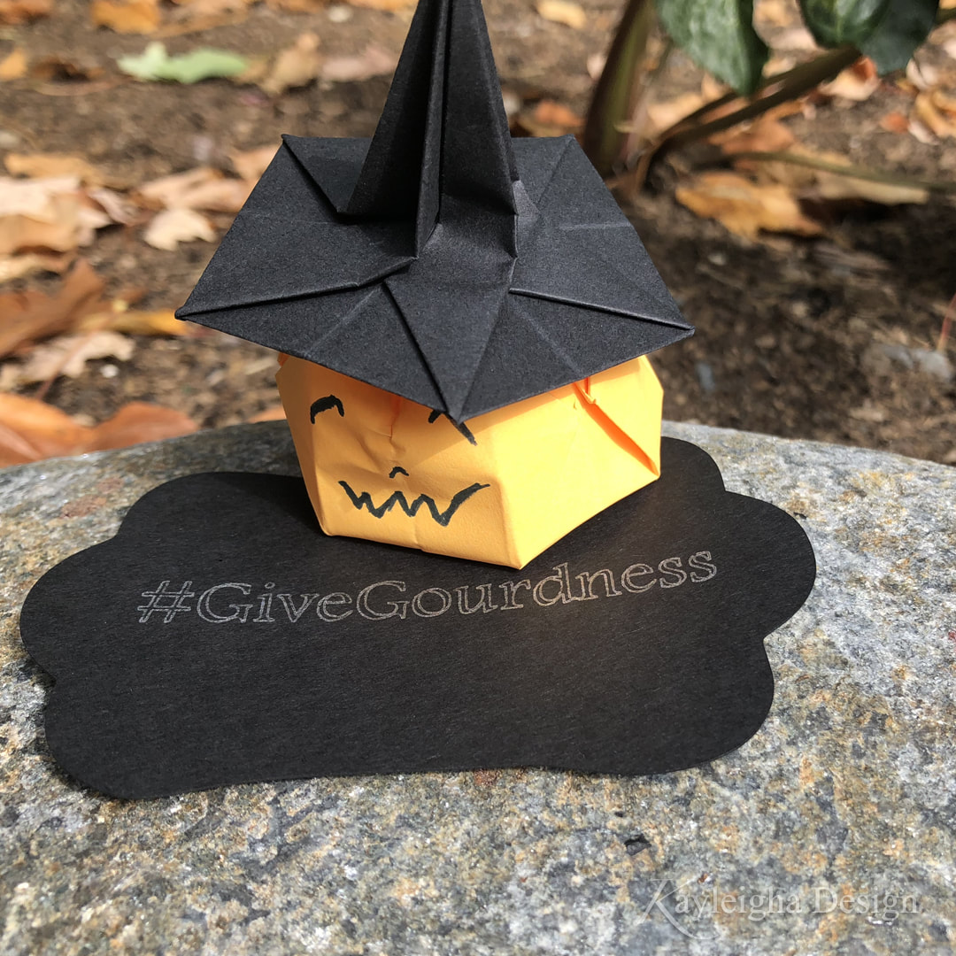 An orange paper origami pumpkin with a black paper witch hat sitting on a black tag that says #GiveGourdness
