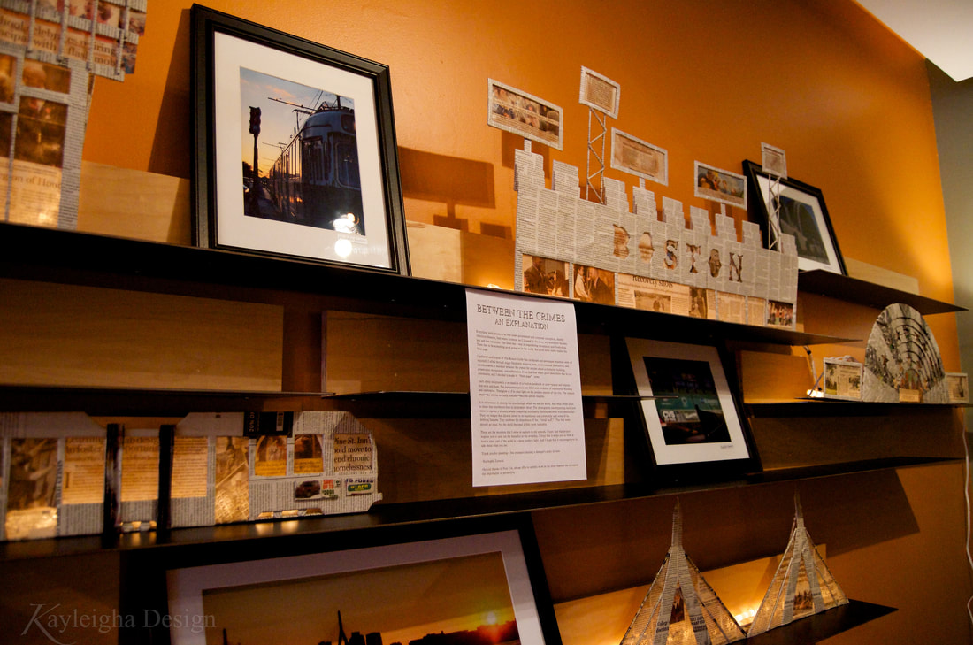 Multiple horizontal shelves contain sculptures made of recycled newspaper. They look like buildings and structures around Boston with transparent areas of newsprint through which warm light shines. Each building has a photograph of a Boston landmark on it. The photos are color photos with white mats and black frames. The wall behind the shelves is orange. 