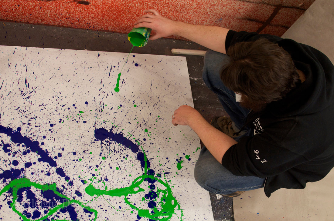 A person with brown hair and wearing a black jacket pours green paint onto a white luan (wood) square. The square is covered in purple and green paint which have been dripped onto it.