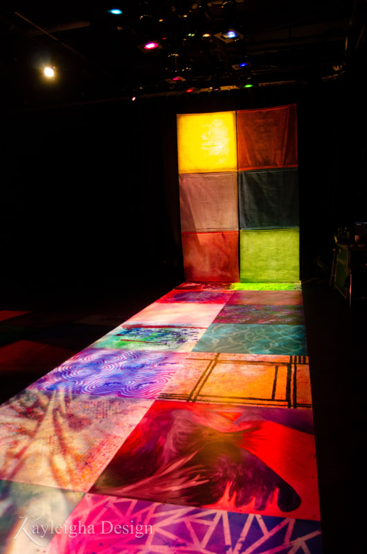 An image of the portal installed. The squares are laid out two by two into a long rectangle. Each is lit in its own box of colorful light. The long rectangle appears to continue up the wall. Six squares of single-color fabric are hung two by two to continue the long rectangle up into the air.