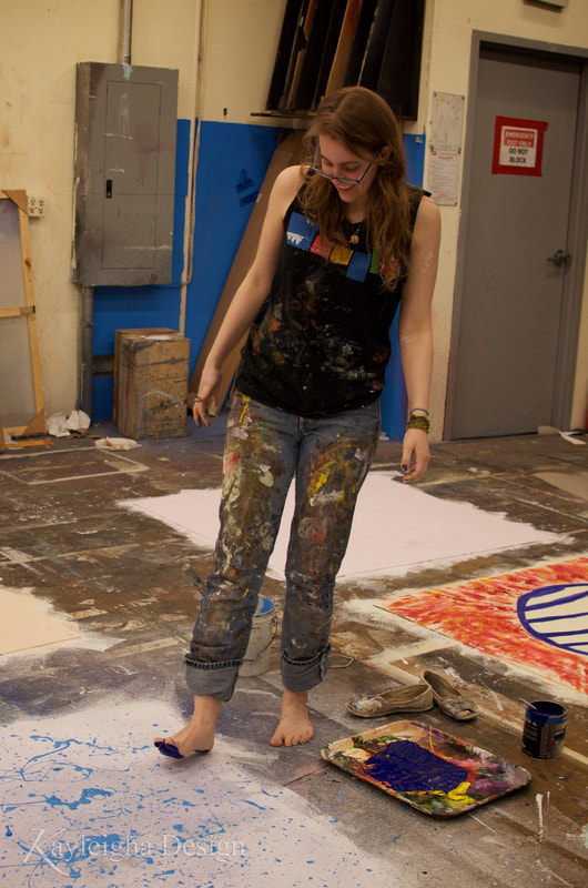 A smiling blonde person with long hair and glasses  and wearing a paint-covered black shirt and jeans steps with a foot covered in paint onto a white square of luan.