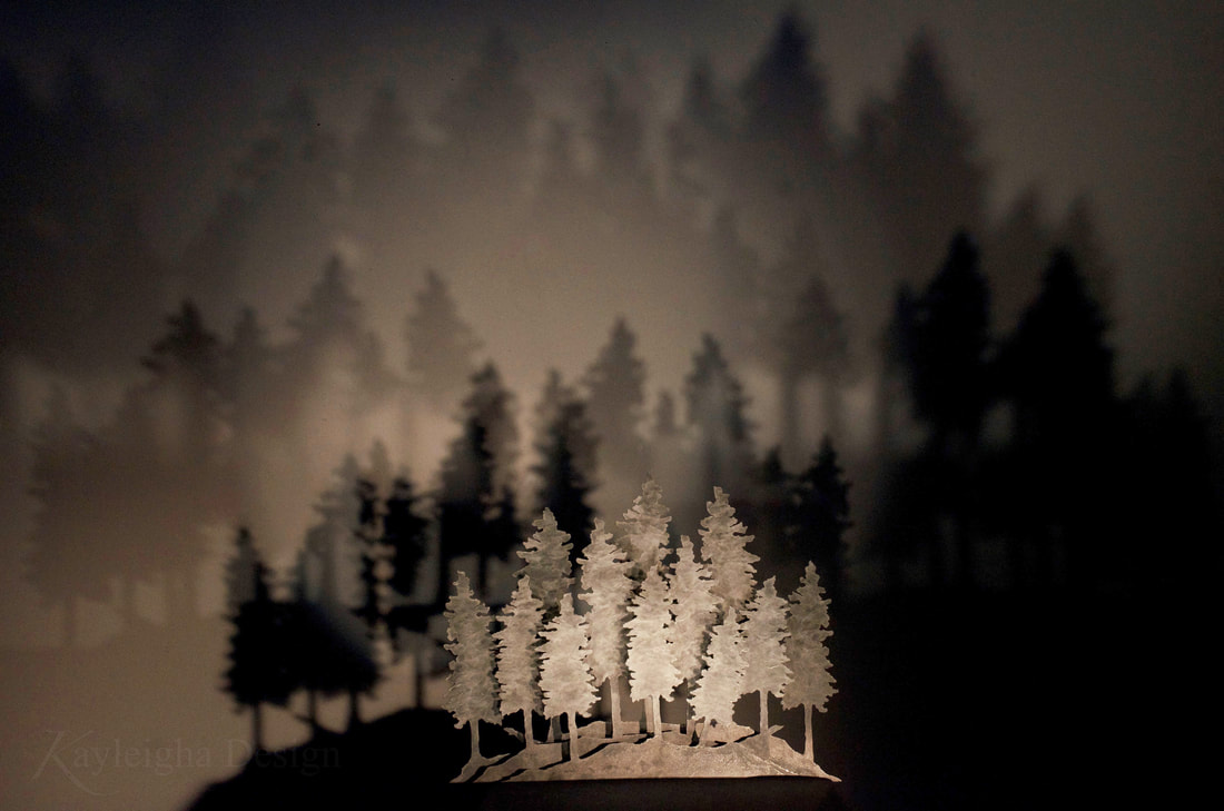 A steel sculpture made of three layers of metal trees cut out of metal and layered one on top of the other. The sculpture is placed in front of a white wall. Light casts shadows of multiple trees onto the wall, creating a light forest.