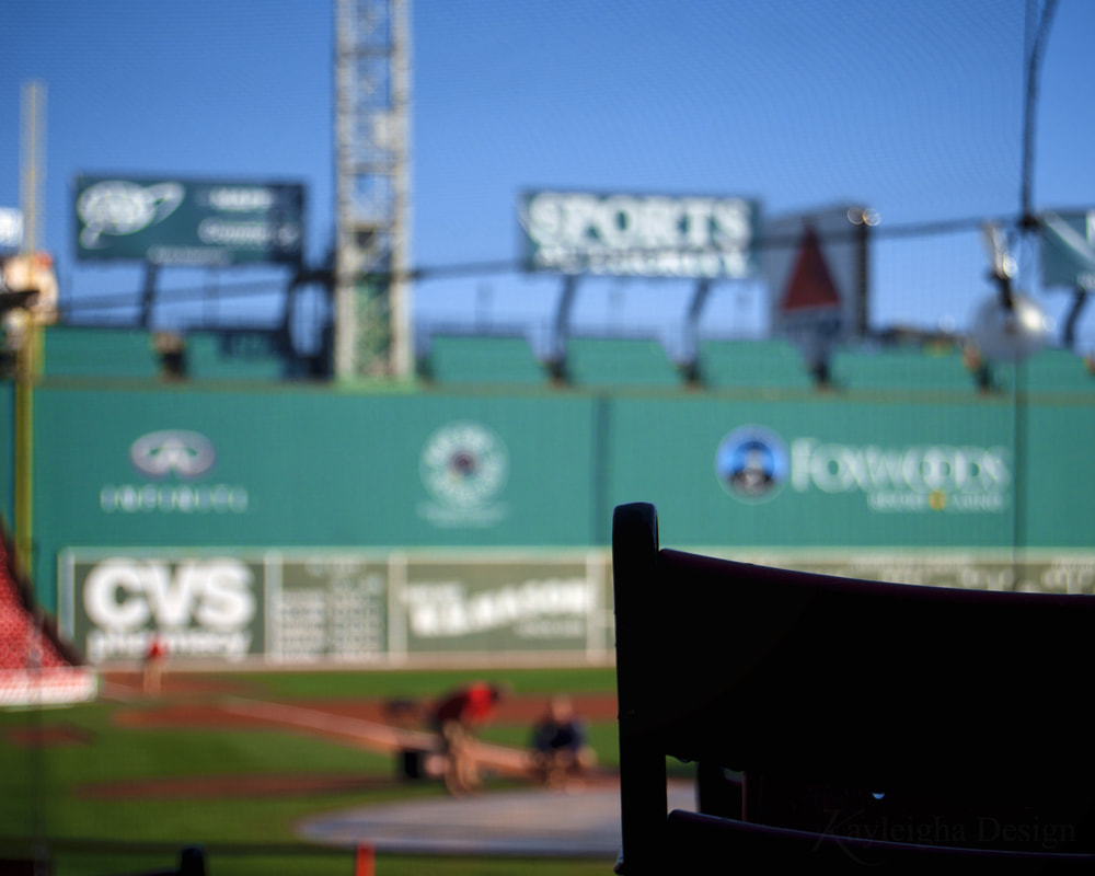 The silhouette of a seat in Fenway Park against the Green Monster, a giant green wall with billboards and lights on trusses above it. The wall is photographed through netting intended to protect people from getting hit by baseballs. Two indistinct figures crouch on the filed in front of the wall. 