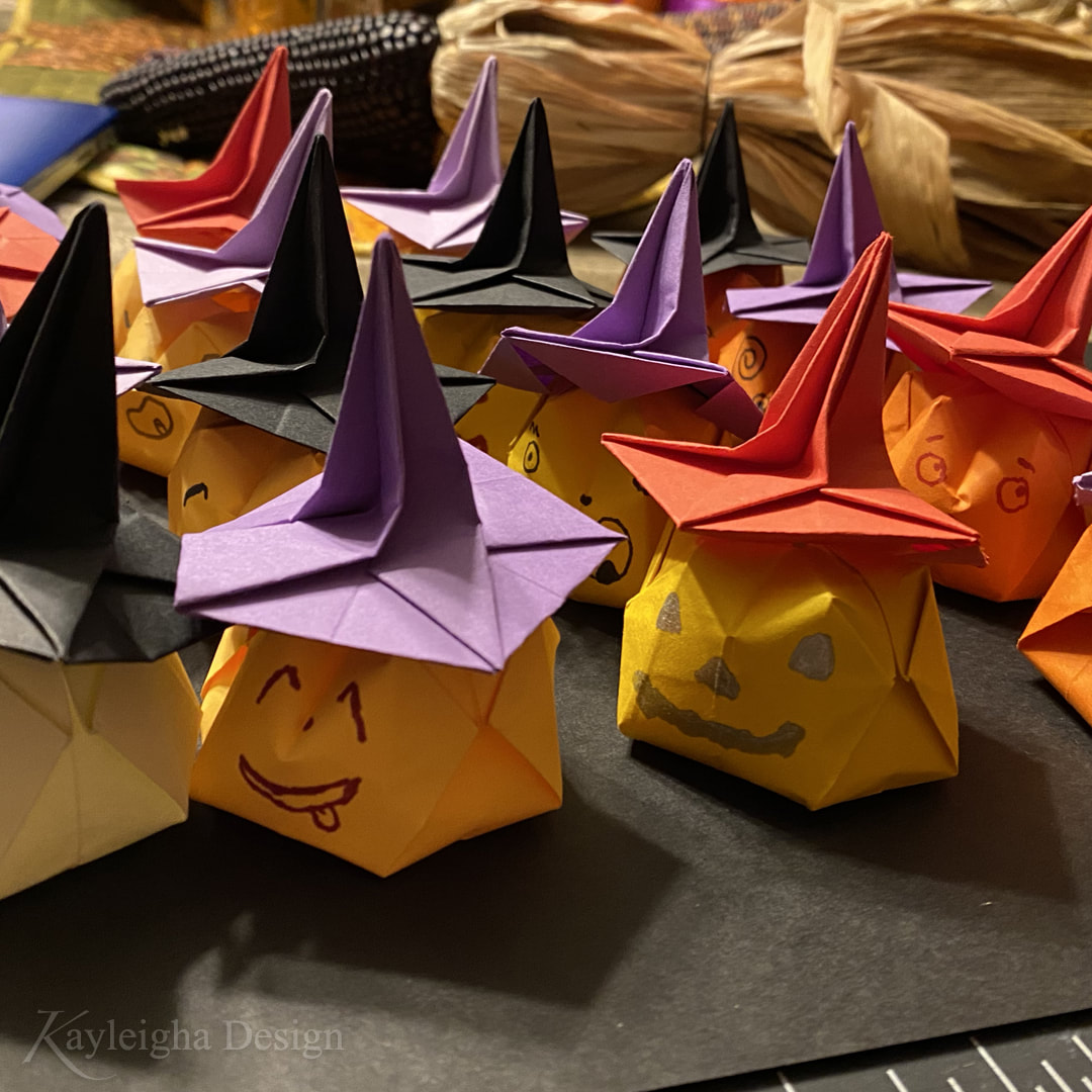An assortment of paper origami pumpkins in oranges, yellows, and whites, wearing red, purple, and black paper witch hats and smiling with drawn-on faces.