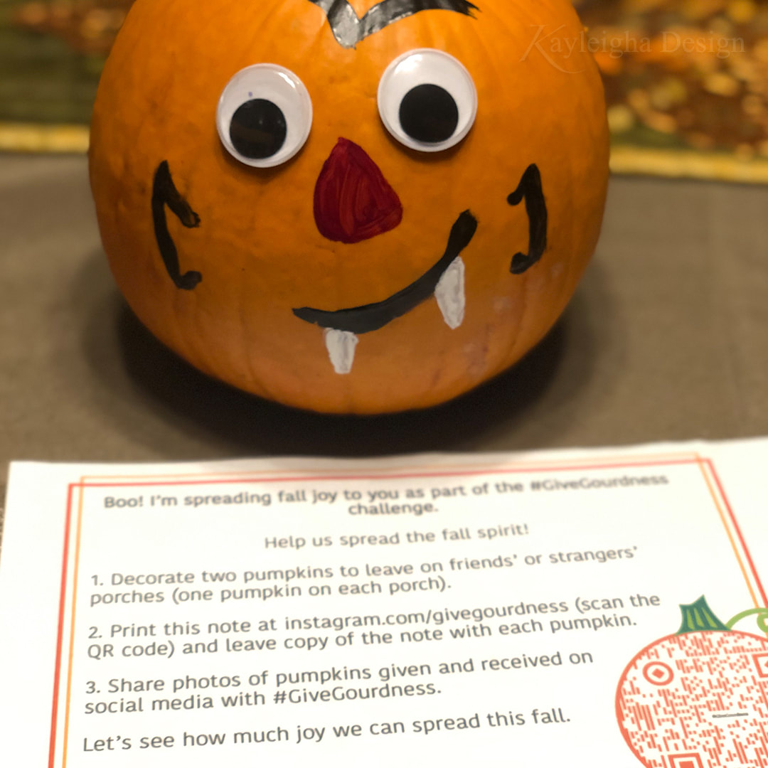 A small orange pumpkin with googley eyes and a painted on vampire face, sitting next to a note encouraging people to leave pumpkins for each other.