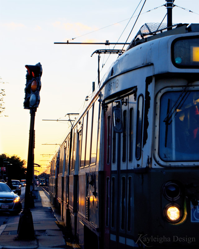 A color photograph of a rusty T subway car. It is white on top and green on the bottom and stopped next to a stop light on its left. It is backlit by the setting sun, causing the train to be bathed in an amber glow.