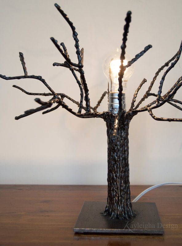 A lamp with a thick square metal base that has a tree growing out of it. There is an old-fashioned light bulb emerging out of the center of the tree at the top, so the the filaments are visible and the light bulb is surrounded by metal branches.