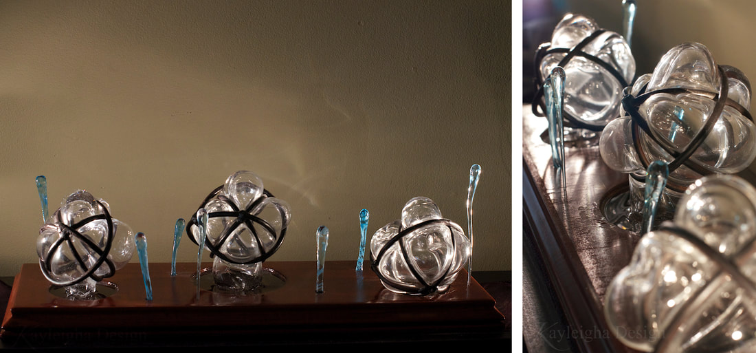 Images of clear glass spheres bubbling out of metal orbs created by welding multiple circles of metal rod together into spheres. There are three of these, resting on a dark-stained wooden base and surrounded by thin blue rods of glass which appear to be dripping, but upwards, out of the wooden base.